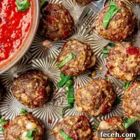 Pizza meatballs on a decorative sheet pan next to a bowl of pizza sauce. Everything is topped with fresh basil and red pepper flakes.