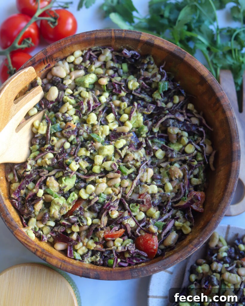 An overhead view of a wooden bowl filled with southwest 3 bean salad made with pintos, white and black beans, corn, tomatoes, fresh cilantro, jalapeno and avocado, with a bowl of lime cilantro salad dressing, some tomatoes fresh cilantro leaves and a plate of salad around the bowl.