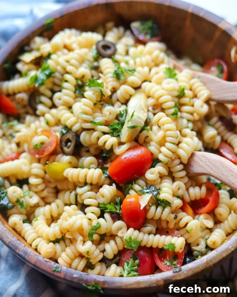 overhead view of a wooden serving bowl with vegan puttanesca pasta salad with lots of fresh tomatoes, olives, artichokes, and herbs.