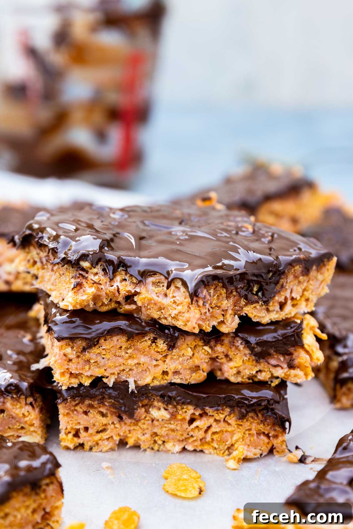 Closeup of 3 bars on top of each other with a bite out of the top one, all on baking paper with more bars at the side and a few cornflakes and with the empty melted chocolate jug in the background.