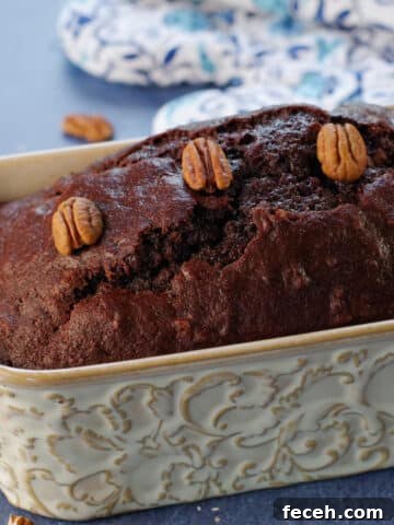 Entire loaf of chocolate zucchini loaf with blue patterned oven mitts in the background and pecans strewn about