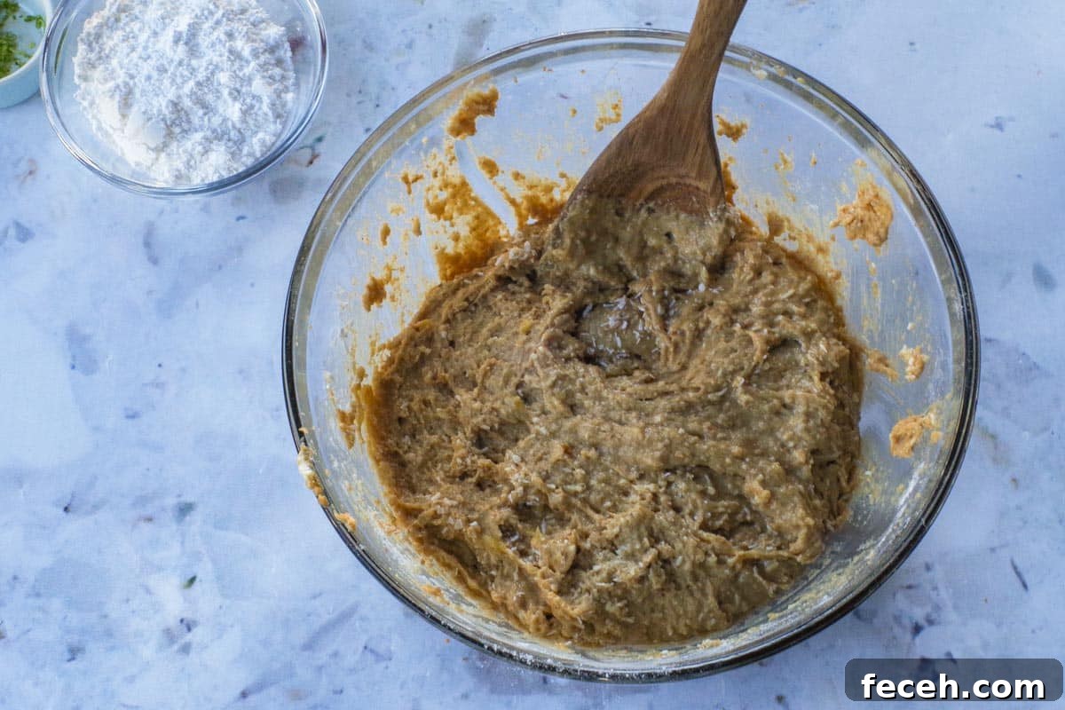 Coconut being stirred into batter with a wooden spoon, in a large glass bowl.