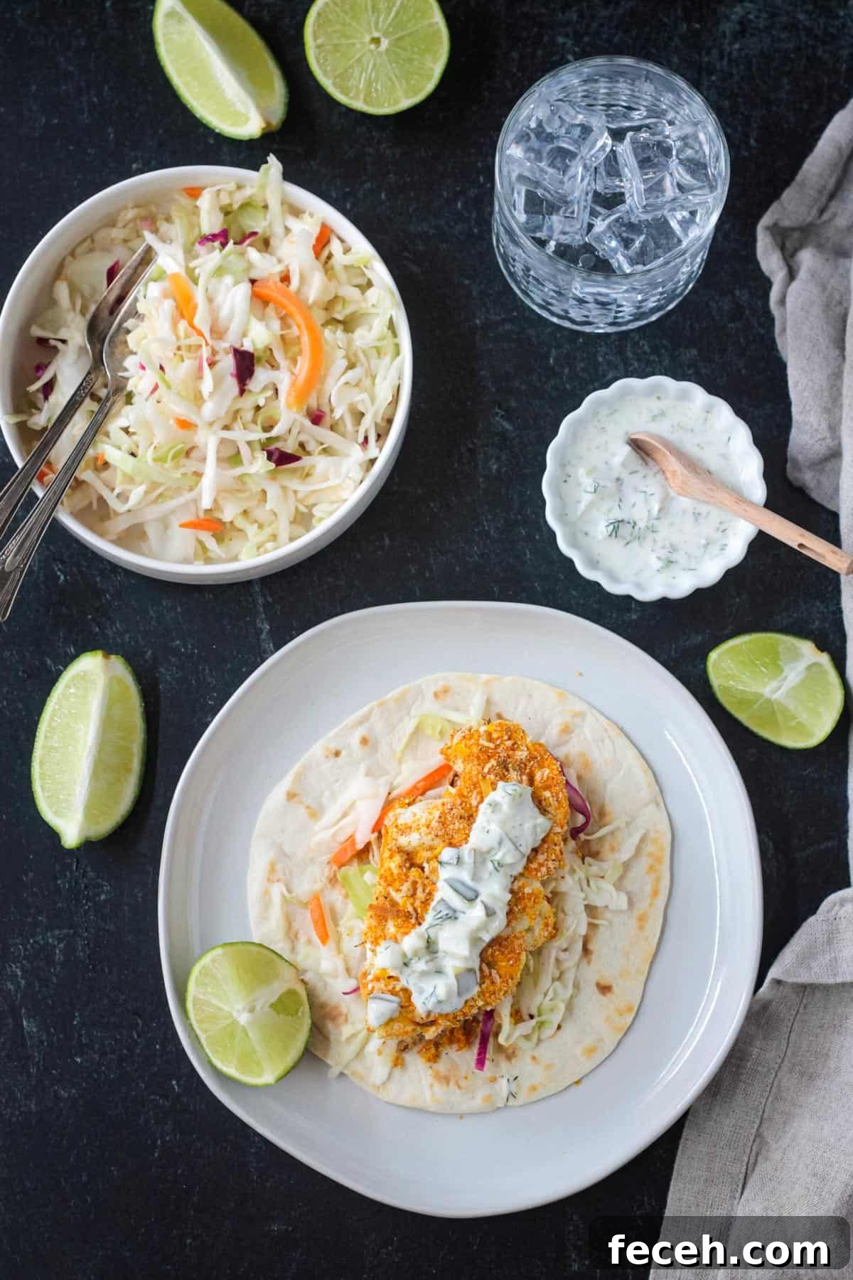 Tablescape with one cauliflower taco on a plate, bowl of coleslaw, bowl of tartar sauce, glass of water, and fresh lime wedges scattered about.