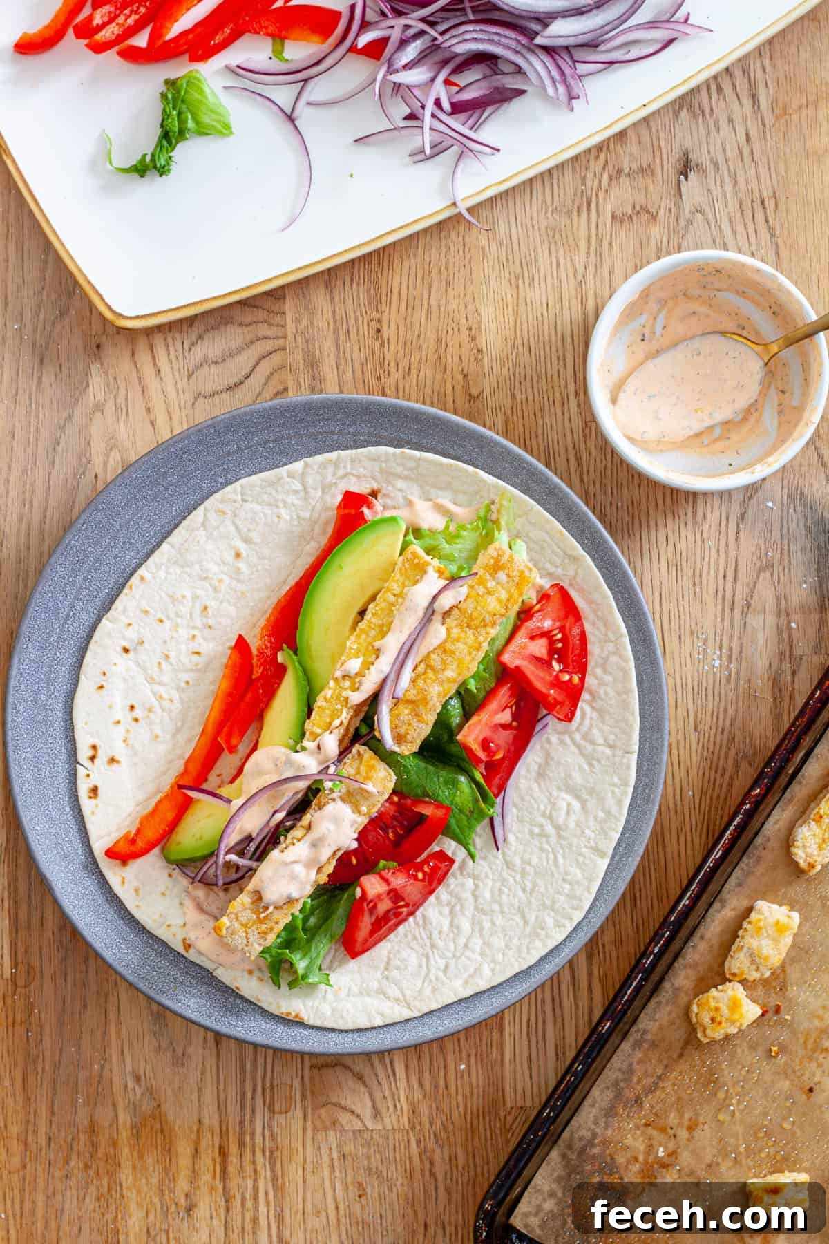 A plate with crispy buffalo tofu wrap topped with avocado, red peppers, lettuce, red onions and a homemade buffalo ranch sauce.