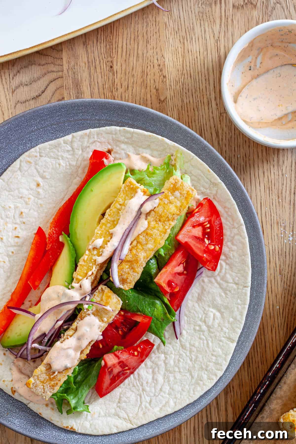 A plate with crispy buffalo tofu wrap topped with avocado, red peppers, lettuce, red onions and a homemade buffalo ranch sauce.
